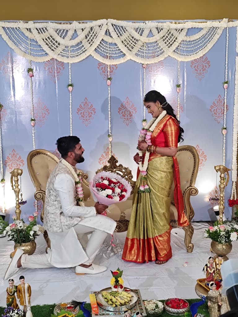 Yathish on one knee offering Rakshitha a bouquet of roses, both wearing jasmine garlands